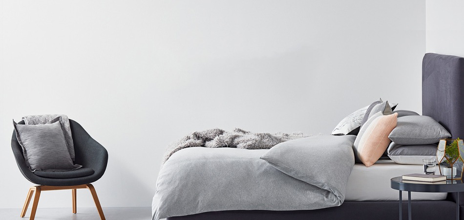 Modern bedroom with a low platform bed, grey bedding, and assorted pillows; dark upholstered headboard and a small round side table on the right.